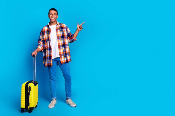 Full size portrait of nice young man suitcase direct finger empty space wear shirt isolated on blue color background. - Photo, image