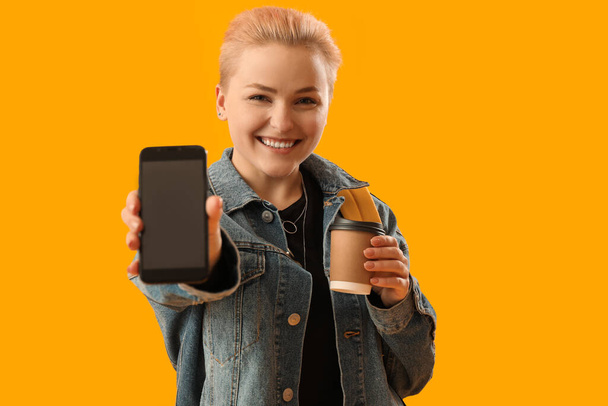 Estudiante femenina con taza de café y teléfono móvil sobre fondo amarillo - Foto, Imagen