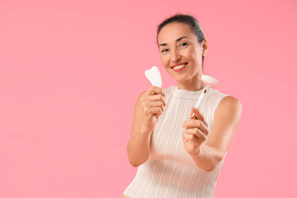 Hermosa mujer madura con herramientas de masaje facial sobre fondo rosa - Foto, Imagen