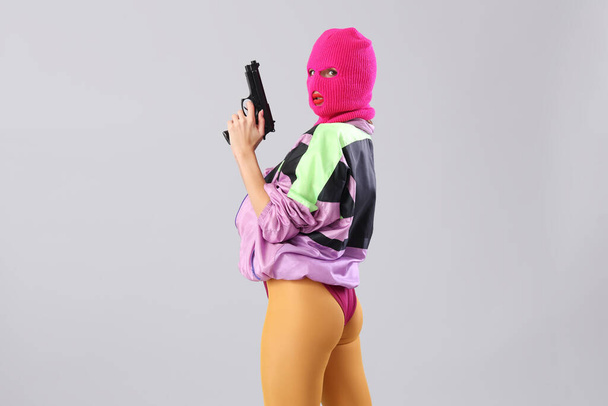 Красивая молодая стильная женщина в балаклаве с пистолетом на сером фоне - Фото, изображение
