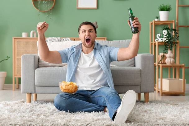 Όμορφος νεαρός άνδρας με μπολ με νόστιμα πατατάκια και μπύρα βλέποντας τηλεόραση στο σπίτι - Φωτογραφία, εικόνα