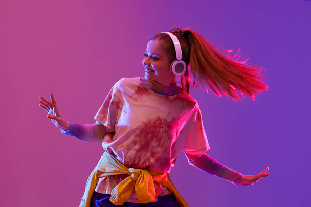 Красивая молодая счастливая женщина в наушниках танцует хип-хоп на темно-фиолетовом фоне - Фото, изображение