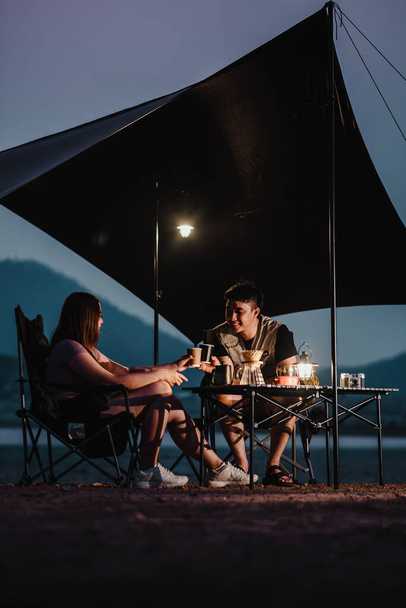 In the serene twilight, a couple shares a warm coffee at a well-equipped campsite, nestled by the lake under a spacious tarp. - Photo, Image