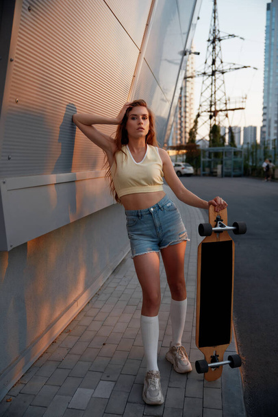 Zwykła młoda kobieta z nowoczesnym longboardem stojąca nad miejskim krajobrazem. Jazda na desce aktywny styl życia - Zdjęcie, obraz