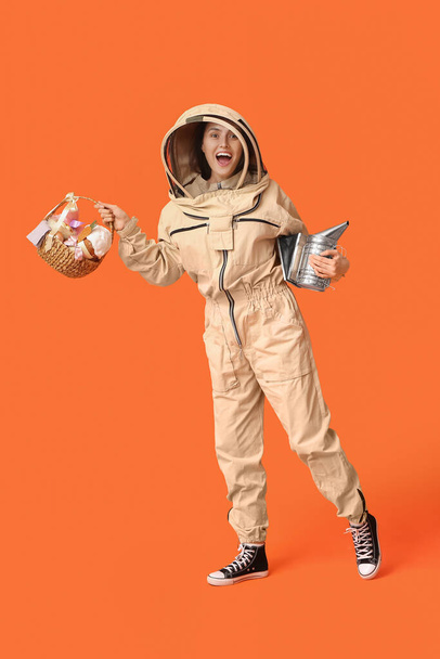 Apicultora feliz en traje protector con cesta de Pascua y fumador sobre fondo naranja - Foto, Imagen