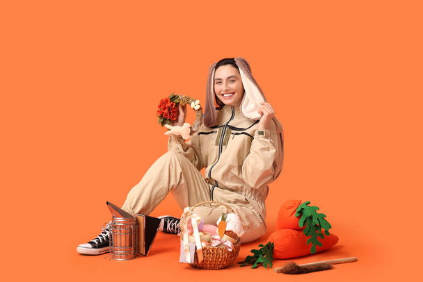 Apicoltore donna felice in orecchie di coniglio con cesto di Pasqua, carote giocattolo e strumenti seduti su sfondo arancione - Foto, immagini