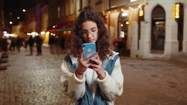 Счастливая улыбчивая брюнетка-подросток в очках с помощью смартфона просматривает текстовые сообщения в социальных сетях. Молодой турист на городской улице ночью. Стиль жизни - Кадры, видео