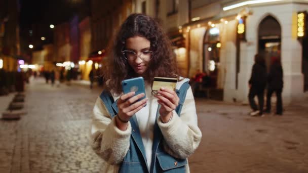 Νεαρή έφηβη κοπέλα που χρησιμοποιεί smartphone πιστωτικών καρτών κατά τη μεταφορά χρημάτων, αγορές online ψώνια, παράδοση τροφίμων παραγγελία, κράτηση δωματίου σε ξενοδοχείο. Ευτυχισμένη γυναίκα σε αστικό δρόμο της πόλης το βράδυ - Πλάνα, βίντεο