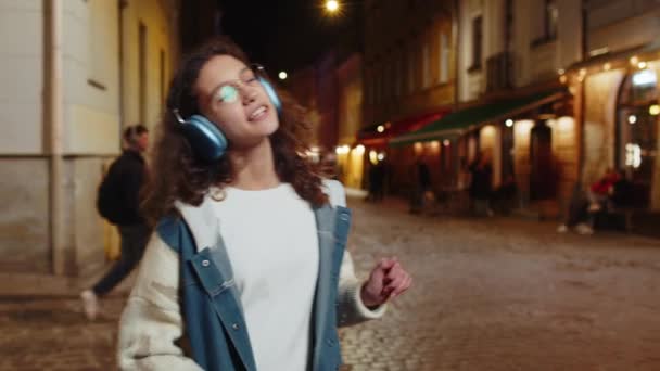 Kablosuz kulaklıklı mutlu, gevşemiş genç kız dışarıda akıllı telefonda dans ederek en sevilen disko rock n roll müziğini dinliyor. Gece şehir caddesinde yürüyen bir turist.. - Video, Çekim