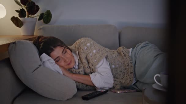Ύπνος γυναίκα lounging καναπέ αναζητούν τηλεόραση στο σπίτι το βράδυ περιβάλλον. Χαλαρή κυρία να αλλάζει κανάλια στον αναπαυτικό καναπέ κάτω από κουβέρτα. Οικιακό μοντέλο χαλαρώνοντας μόνο του στον ύπνο στο φως λάμπα ατμόσφαιρα - Πλάνα, βίντεο