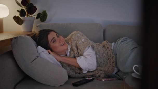Mosolygó barna kapcsoló TV csatornák pihentető kanapé késő esti közelkép. Boldog nő, aki egyedül nézi a komédiát. Pozitív örömteli lány pihenő nappali szundikáló hangulatos lakás hely - Felvétel, videó