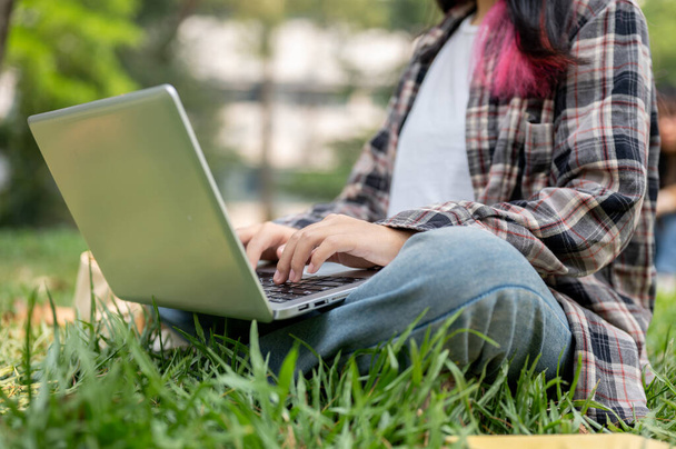 Rajattu kuva naispuolinen opiskelija rento kulumista käyttää hänen kannettavan tietokoneen istuessaan nurmikolla puistossa aurinkoisena päivänä. langattoman teknologian käsite - Valokuva, kuva