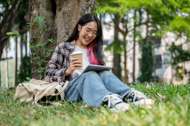 Μια νεαρή Ασιάτισσα με καθημερινά ρούχα κάθεται κάτω από ένα δέντρο σε ένα πάρκο της πανεπιστημιούπολης, κρατώντας ένα φλιτζάνι καφέ και μια ταμπλέτα, απολαμβάνοντας μια ηλιόλουστη μέρα, ενώ σπουδάζει σε εξωτερικούς χώρους. έννοιες πανεπιστημιακής ζωής και ασύρματης τεχνολογίας - Φωτογραφία, εικόνα