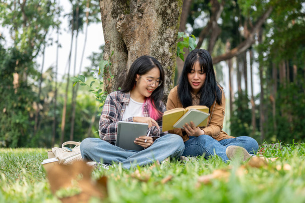 Δύο νεαρές Ασιάτισσες φοιτήτριες κάθονται στο γρασίδι κάτω από ένα δέντρο, μιλώντας και διαβάζοντας μαζί σε ένα πάρκο της πανεπιστημιούπολης μια ηλιόλουστη μέρα. φιλία και πανεπιστημιακή ζωή έννοιες - Φωτογραφία, εικόνα