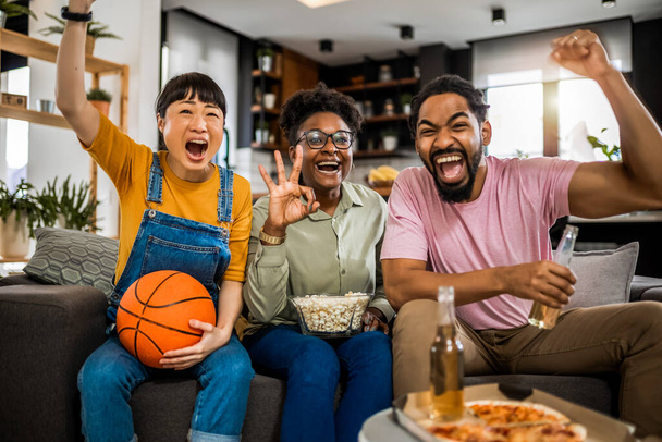 Multiraciální skupina přátel, kteří sledují basketbal, jedí pizzu, pijí pivo a jásají. - Fotografie, Obrázek