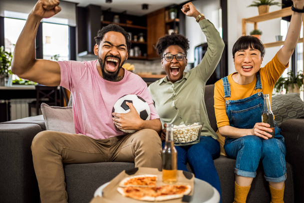 Eine multiethnische Gruppe von Freunden schaut Fußballspiele, jubelt, isst Pizza und trinkt Bier. - Foto, Bild
