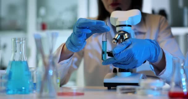 Wissenschaftler in Handschuhen halten und kippen im Labor ein Reagenzglas mit einer blauen Flüssigkeit und einem Mikroskop. Giftige Forschung - Filmmaterial, Video