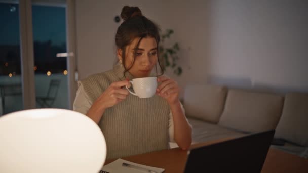 Večer na volné noze pití kávy šálek vypadá notebook obrazovku na pohodlné domácí pracoviště detailní up. Vážná žena, která se dívá na webinární video, studuje klidně sama. Bruneta zaostřená dáma pracuje na lampě - Záběry, video