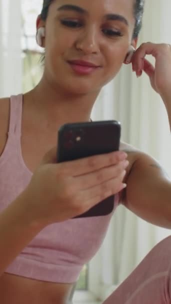 Κατακόρυφη κλίση της θετικής γυναίκας που φοράει ακουστικά ενώ χρησιμοποιεί κινητό τηλέφωνο μετά από έντονη προπόνηση στο σπίτι - Πλάνα, βίντεο