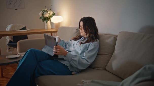 Evening girl watching pad screen at comfy home closeup. Relaxed woman looking tablet video relaxing calmly alone. Brunette lady sipping tea sitting cozy sofa at lamp light interior. Domestic routine - Footage, Video