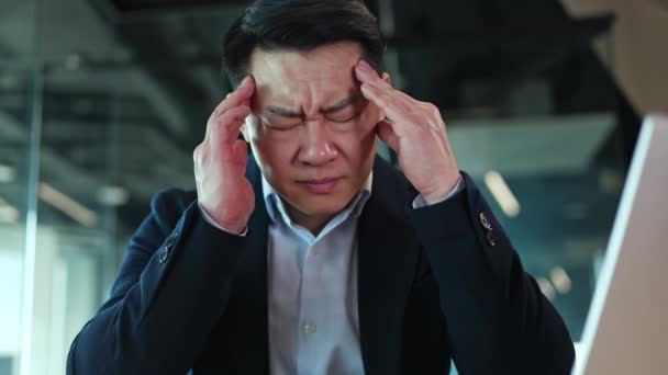Detailní záběr asijského muže oblečeného v klasickém obleku masírujícím spánky s oběma rukama a zároveň pocit akutní bolesti hlavy. Vyčerpaný kancelářský zaměstnanec trpící nepříjemnými příznaky z důvodu stresující práce. - Záběry, video