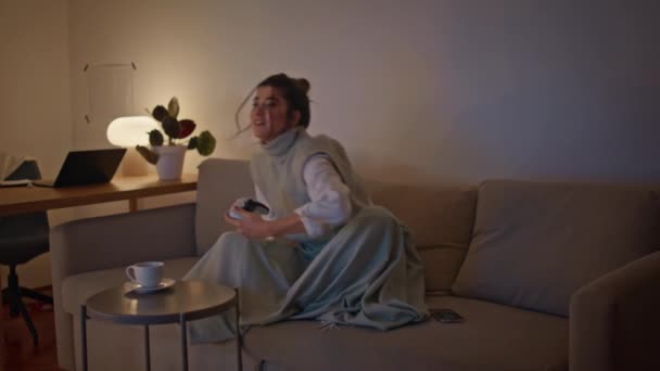 Χαμογελώντας gamer κρατώντας joystick παίζει video game στο σπίτι. Χαρούμενη γυναίκα που πίνει τσάι χαλαρώνοντας το βράδυ στο σαλόνι. Ενθουσιασμένη κυρία που χρησιμοποιεί χειριστήριο στο άνετο διαμέρισμα αργά. Έννοια ψυχαγωγίας - Πλάνα, βίντεο