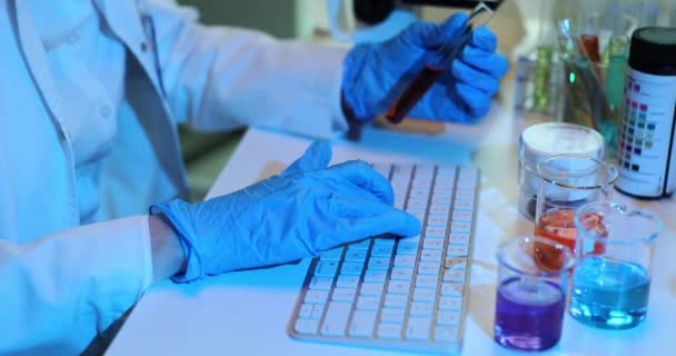 Científico en guantes azules utiliza una computadora en un laboratorio y sostiene un tubo de ensayo con sangre o líquido tóxico oscuro. Químico que usa computadora para registrar datos mientras sostiene el tubo de ensayo con líquido rojo - Imágenes, Vídeo