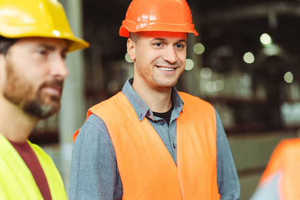 Attractive, smiling man, worker, foreman wearing hard hat talking with colleagues, listening while standing in warehouse. Concept of industry, cooperation, communication - Photo, Image