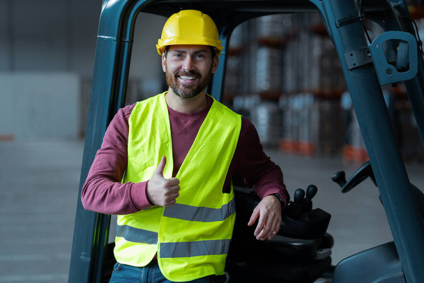 Attractive, handsome, bearded man, driver showing thumbs up, wearing helmet, standing near forklift, looking at camera, standing in warehouse. Concept of logistics, vehicle - Photo, Image