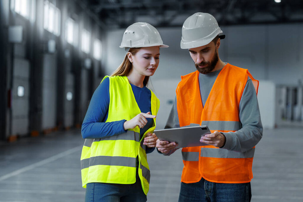 Attractive man and woman, engineers wearing protective workwear and hard hat using digital tablet, planning project, standing in warehouse. Online technology concept, teamwork - Photo, Image