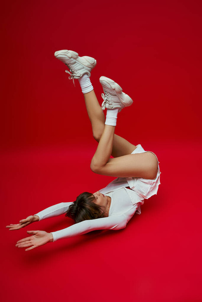 Una mujer con estilo en un traje blanco y zapatos de tenis golpea una pose sobre un vibrante telón de fondo rojo. - Foto, imagen