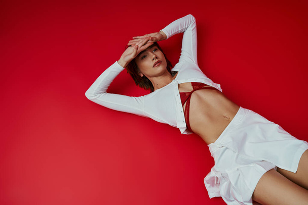 Een stijlvolle vrouw in een wit shirt en ligt sierlijk op een levendige rode vloer. - Foto, afbeelding