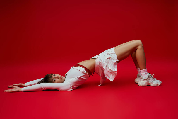 Stylowa młoda kobieta w białym ubraniu leżąca z wdziękiem na ziemi na tętniącym życiem czerwonym tle. - Zdjęcie, obraz