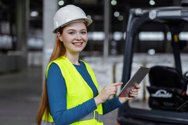 Beautiful, cheerful, young woman, successful engineer wearing hard hat, using digital tablet, looking at camera. Attractive female worker working in warehouse. Online technology concept - Photo, Image