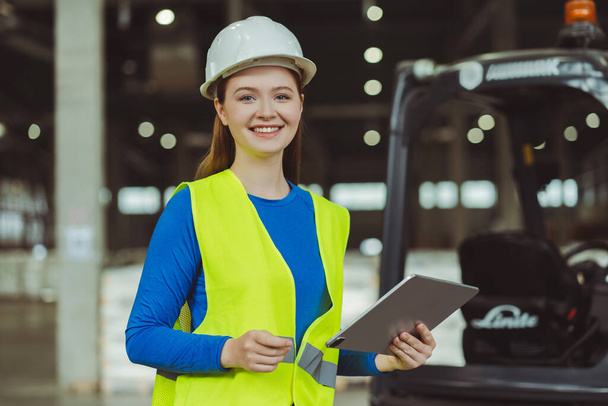 Attractive smiling young woman, successful engineer wearing hard hat, using digital tablet, looking at camera. Female worker working in warehouse. Online technology concept - Photo, Image