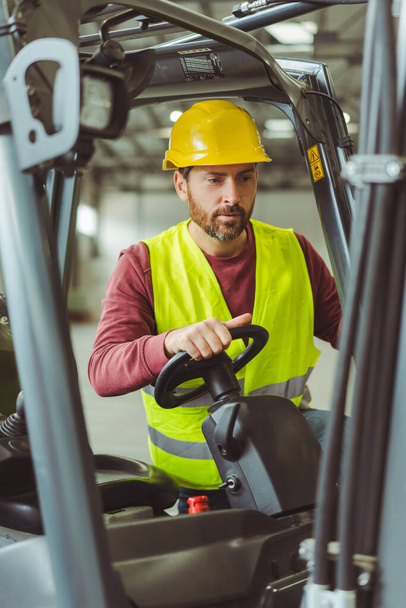 Attractive bearded man wearing yellow protective helmet and workwear driving forklift, working in warehouse. Concept of transportation, vehicle - Photo, Image