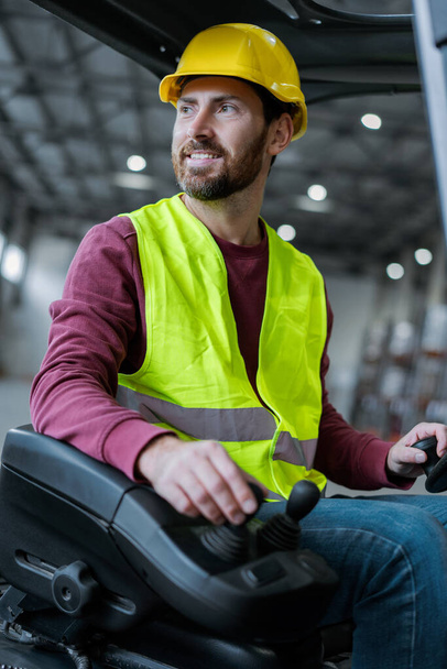 Smiling, handsome worker, professional driver wearing hard hat and workwear driving forklift looking away, working in warehouse. Concept of logistics, equipment, transportation - Photo, Image