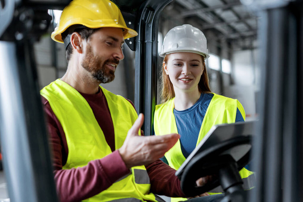 Handsome colleagues in helmets and communicate with each other, man driving forklift, using digital tablet, mobile app working in warehouse. Concept of transportation, cooperation, communication - Photo, Image