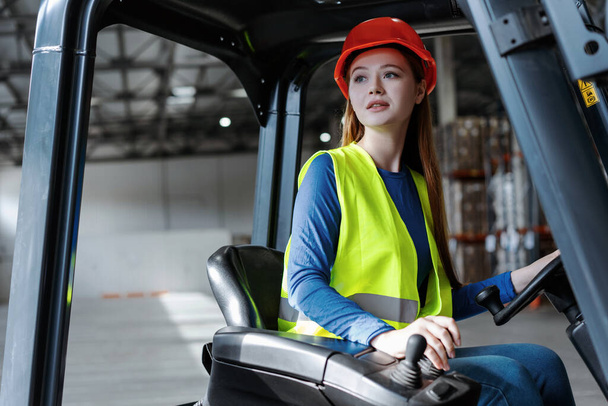 Beautiful, cute female worker, young woman wearing hard hat and work clothes driving forklift, looking away. Attractive engineer driving warehouse. Concept of logistics, transportation, vehicle - Photo, Image