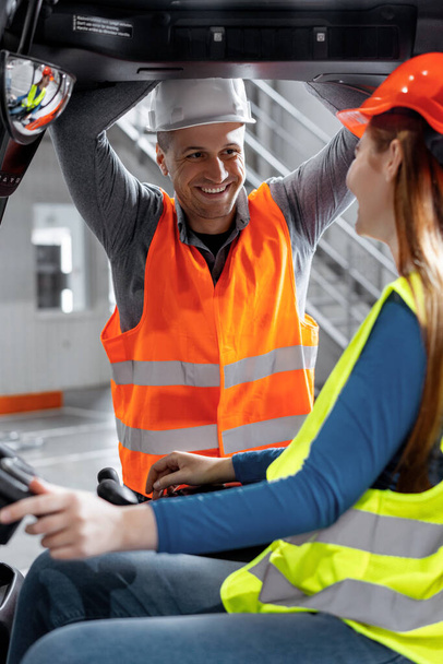 Attractive colleagues wearing hard hats and work clothes, young woman driving forklift, talking to foreman at work in warehouse. Industry concept, vehicle - Photo, Image