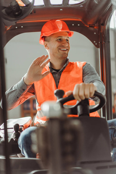 Handsome, smiling man, foreman sitting on forklift, waving his hand, looking away, greeting, wearing hard hat and workwear. Concept of warehouse work, transportation, vehicle - Photo, Image