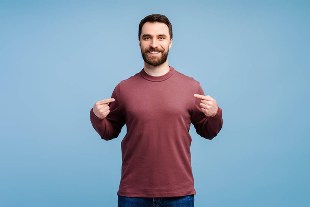 Sonriente hombre barbudo guapo, moderno hipster con camisa en blanco apuntándose aislado sobre fondo azul. Burla. - Foto, imagen