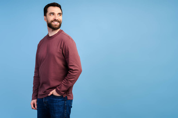 Портрет красивого улыбающегося бородатого мужчины в осеннем свитере, стильные джинсы на синем фоне. Портрет успешной модели среднего возраста, позирующей для фотографий, студийный снимок - Фото, изображение
