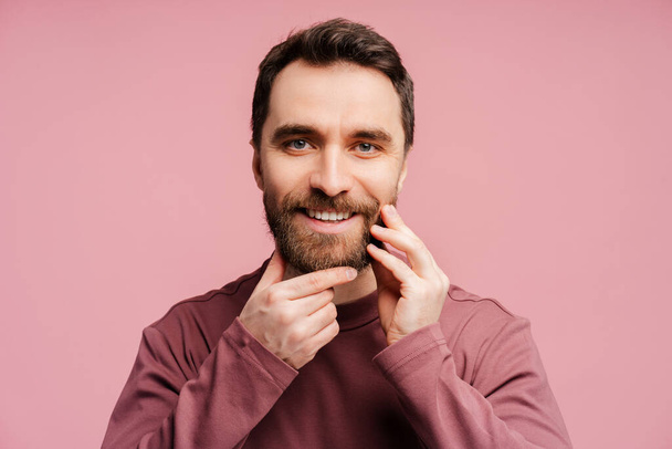 Primer plano retrato de hombre sonriente guapo tocando su cara, barba correcta mirando espejo aislado sobre fondo rosa. Cuidado de la piel, higiene, concepto de rutina matutina - Foto, imagen