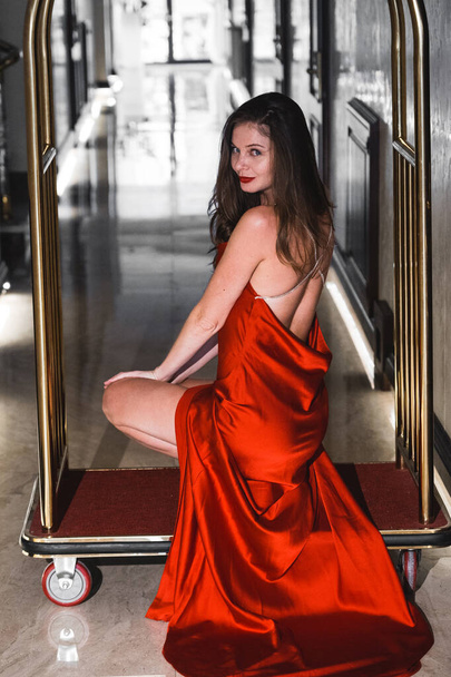 Two sexy women in red dresses ride on a golden luggage trolley in a hotel. Fashion shooting concept - Photo, Image