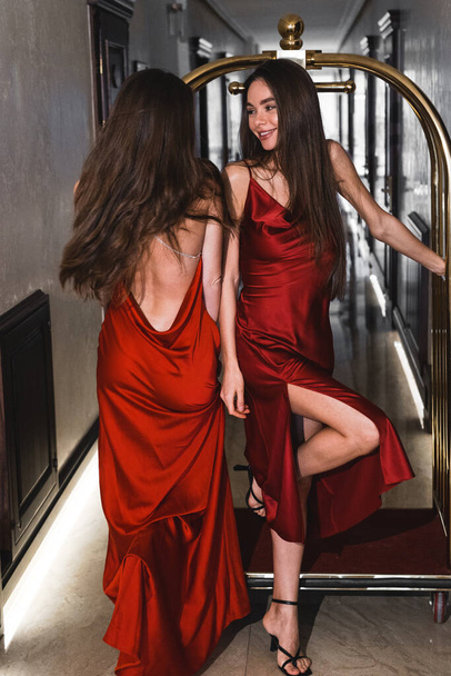 Δύο σέξι γυναίκες με κόκκινα φορέματα οδηγούν σε ένα χρυσό καροτσάκι αποσκευών σε ένα ξενοδοχείο. Έννοια λήψης μόδας - Φωτογραφία, εικόνα