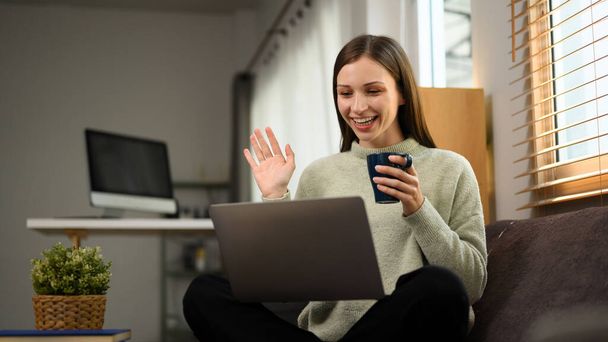 Fröhliche junge Frau winkt mit der Hand und macht Videoanruf am heimischen Laptop. - Foto, Bild