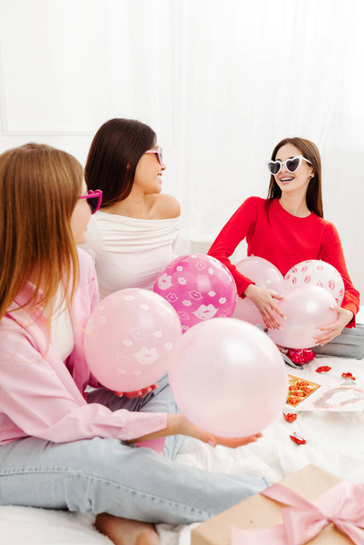 Vista vertical das mulheres bonitas felizes vestindo óculos de sol elegantes sentados na cama com balões e olhando uns para os outros. Jovens modelos de moda atraentes posando para fotos - Foto, Imagem