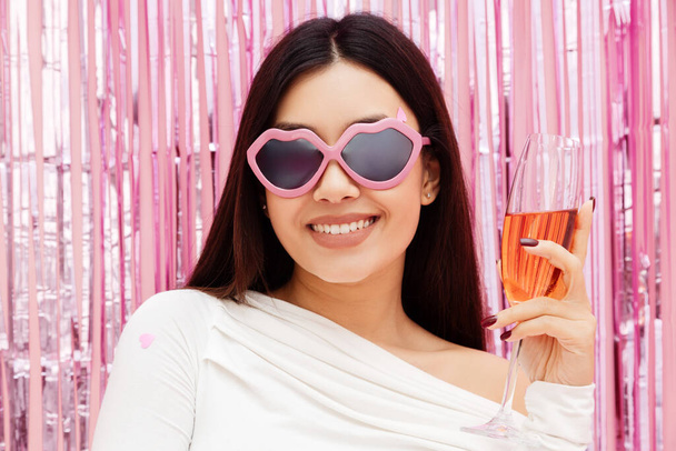 Χαμογελώντας όμορφη γυναίκα φορώντας γυαλιά ηλίου κρατώντας ποτήρι κρασιού, κατά τη διάρκεια γιορτή κόμμα απομονώνονται σε ροζ φόντο. Έννοια γενεθλίων - Φωτογραφία, εικόνα