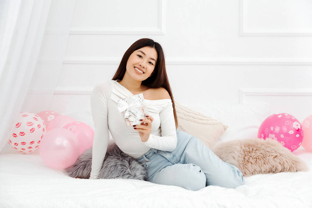 Güzel, gülümseyen Asyalı bir kadın doğum günü hediyesini tutuyor ve rahat bir dairede yatakta oturuyor. Tatil kutlamaları konsepti - Fotoğraf, Görsel